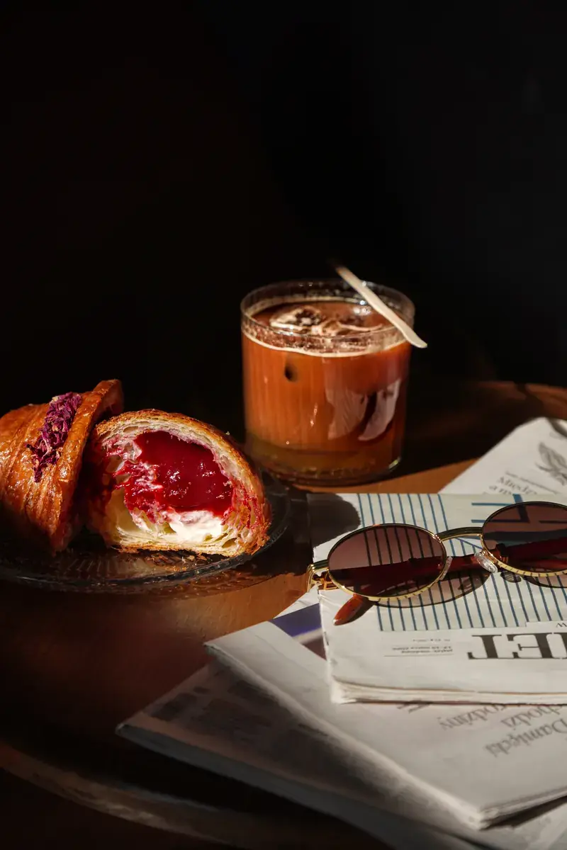 A croissant cut in half with a raspberry filling, next to an iced coffee, sunglasses and a newspaper