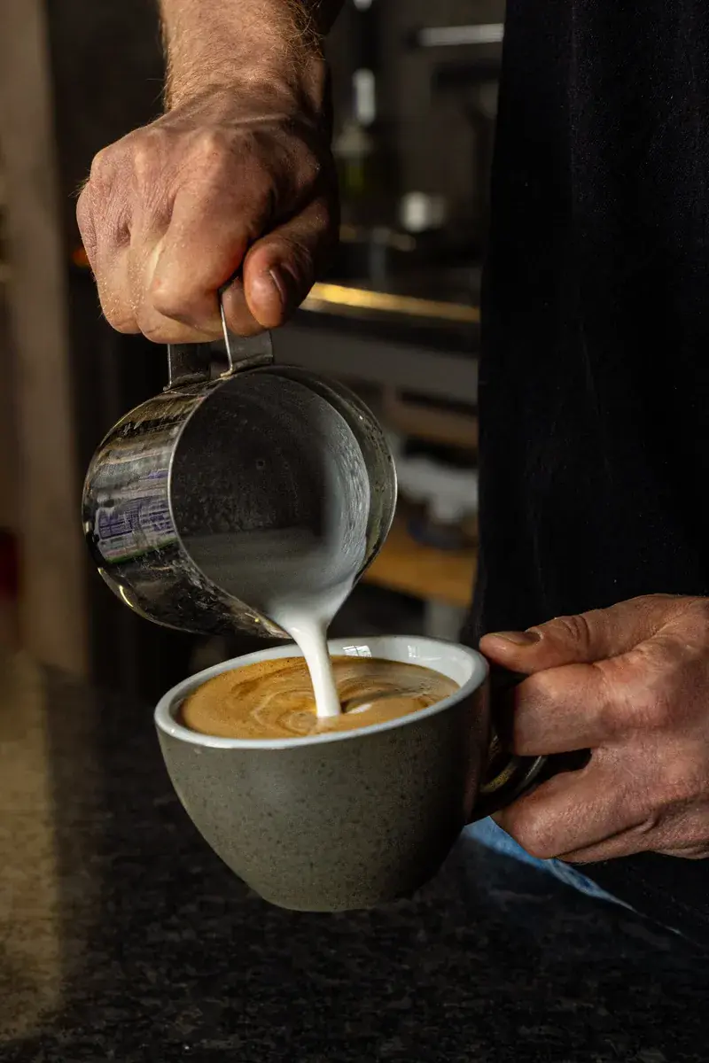 Frothed milk poured from a jug into coffee in a grey cup