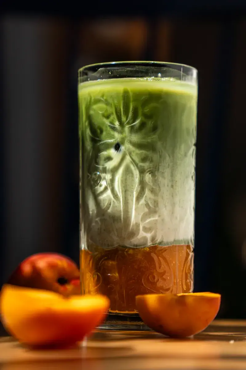 Green matcha in a tall cut-glass tumbler over an orange layer, next to halved peaches