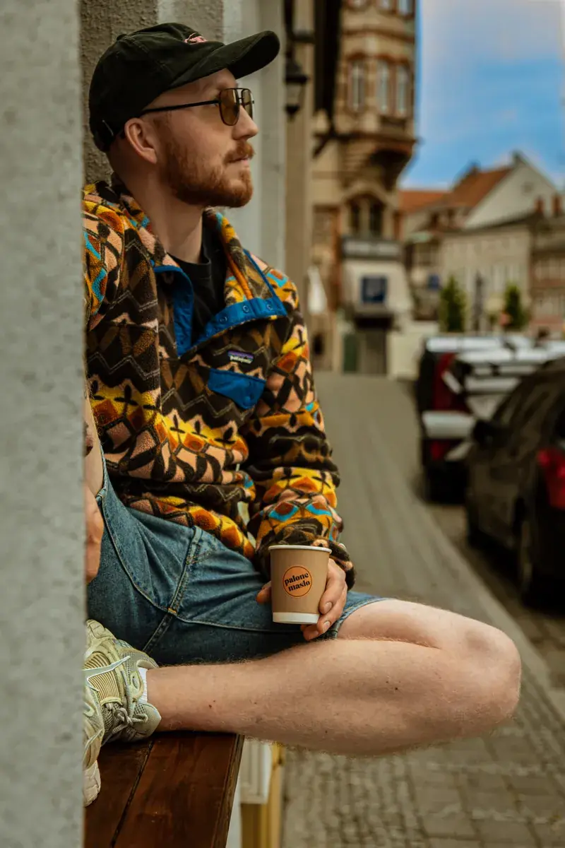 A guest in a cap sitting sideways on a windowsill with a cup of coffee, a street of old townhouses behind