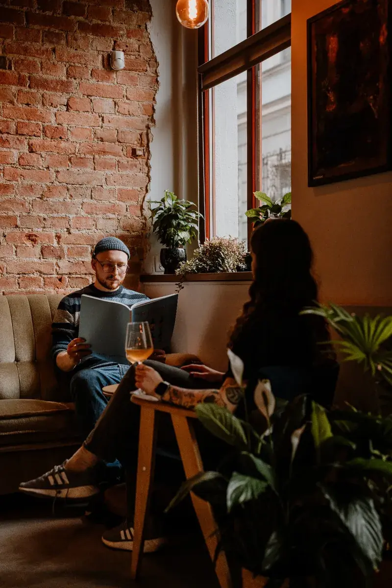 Inside by the brick wall: a guest reading at a table with a glass, next to a big window and plants