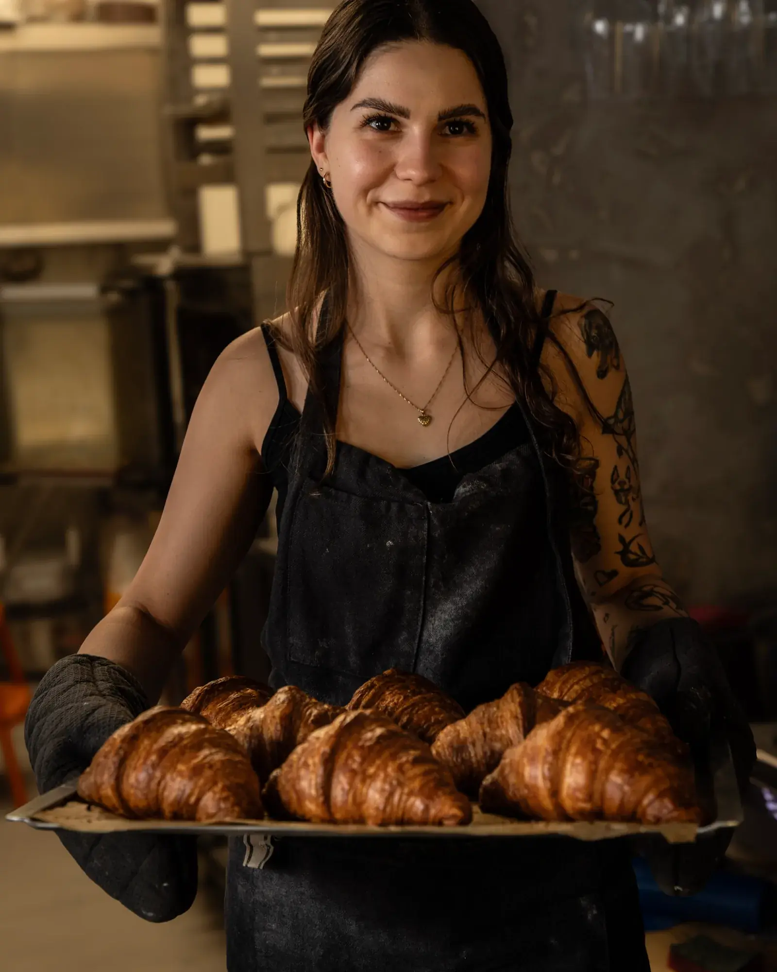 A smiling person in an apron and oven gloves holding a tray of freshly baked croissants