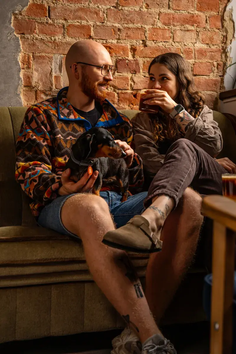 Two guests on a sofa by a brick wall, one holding a dachshund, the other with a glass of iced coffee