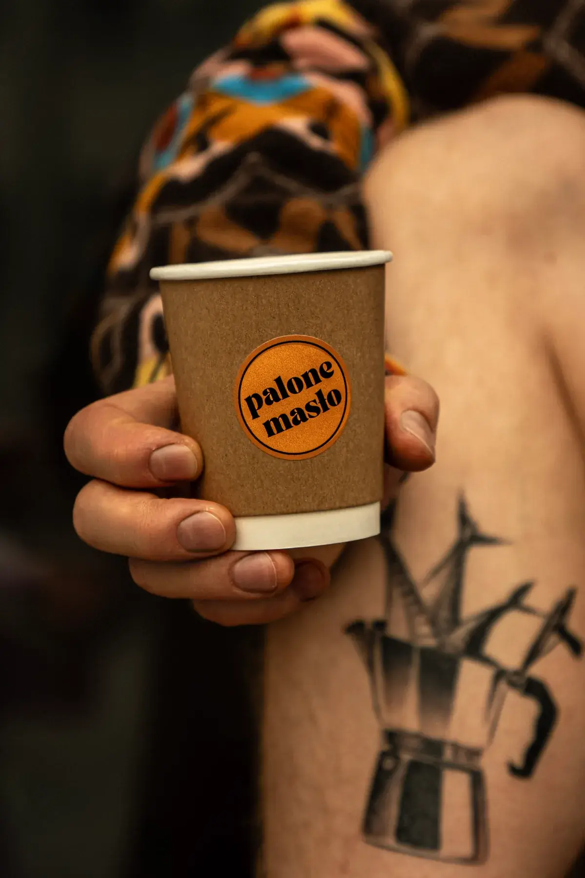 A hand holding a paper takeaway cup with an orange Palone Masło sticker, next to a forearm with a moka pot tattoo