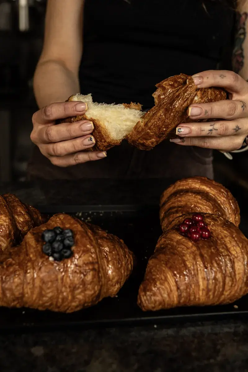 Hands tearing a croissant apart over a tray full of croissants, some topped with fruit